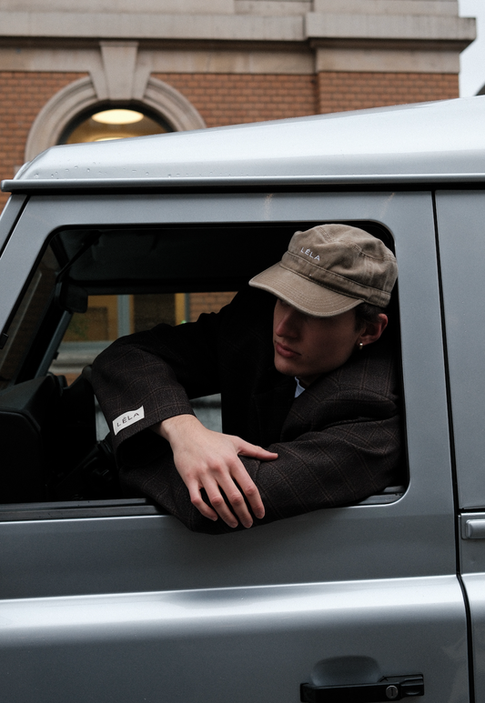 Man wearing washed army green LÉLA cap leaning out of silver car window, styled with brown LÉLA Archer Blazer featuring LÉLA patch