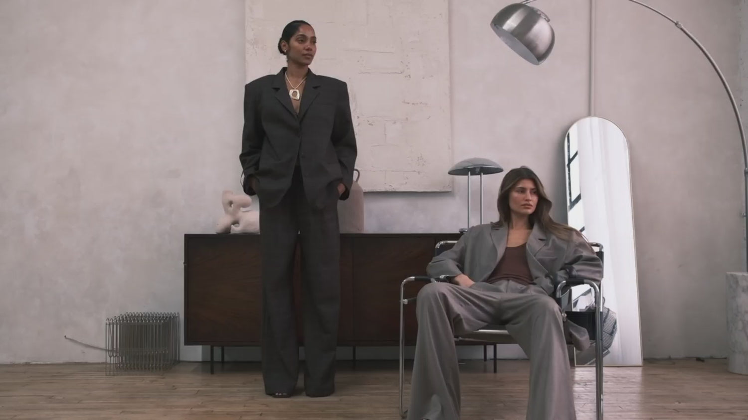 Two models posing in a minimalist studio with concrete walls. One stands wearing an oversized dark gray suit, while the other sits in a chrome chair wearing a light gray suit. Both models stand up and walk out of frame to the left, displaying the draped fit and movement of the LÉLA suits